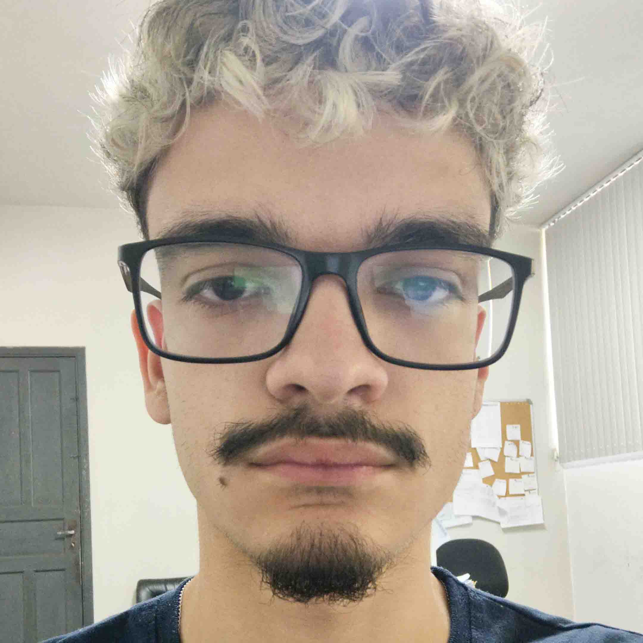 Pedro Mariano, aluno de Engenharia de Produção, posando em um ambiente interno com um fundo claro. Ele usa óculos de armação preta e tem barba e bigode, com cabelos loiros e cacheados e uma expressão neutra.