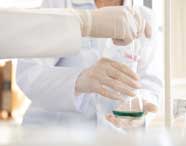 Mãos com luvas de látex manipulam um frasco Erlenmeyer contendo líquido verde, utilizando uma pipeta. A cena ocorre em laboratório do curso de Engenharia de Produção, destacando práticas de análise química e manipulação segura de amostras em ambiente acadêmico.