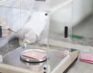 Pessoa com jaleco branco e luvas manuseia espátula metálica para pesar amostra de pó claro sobre papel rosa, dentro de balança analítica de precisão. O procedimento faz parte das análises laboratoriais realizadas por estudantes de Engenharia de Produção.