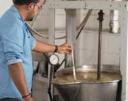 Pessoa com camisa azul opera grande misturador industrial de aço inox, monitorando temperatura com termômetro em tanque cheio de líquido. Ambiente típico do Centro Tecnológico de Alimentos, utilizado por estudantes de Engenharia de Produção para práticas industriais.