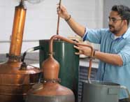 Pessoa com camisa azul manuseia equipamentos de destilação em cobre e ferro no laboratório do Centro Tecnológico de Alimentos. O ambiente apresenta diversos recipientes conectados por tubos, utilizados em processos produtivos do curso de Engenharia de Produção.