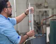 Pessoa com camisa azul segura proveta cilíndrica transparente cheia de líquido, observando medidor interno. Ao fundo, equipamentos industriais e bancada de laboratório, cenário típico de atividades práticas em Engenharia de Produção na área de alimentos.