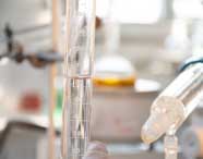 Close em proveta graduada transparente preenchida até a marca de 70 ml, ao lado de outros instrumentos de vidro. Fundo desfocado mostra balão volumétrico com líquido amarelo, compondo ambiente de laboratório químico utilizado por estudantes de Engenharia de Produção.