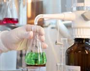 Mão enluvada segura frasco Erlenmeyer com líquido verde sob torneira de equipamento de laboratório. Ao fundo, frascos com líquidos coloridos, incluindo vermelho e rosa, evidenciando práticas de química do curso de Engenharia de Produção.