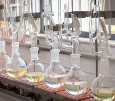 Conjunto de balões volumétricos de vidro conectados a tubos e presilhas metálicas, alinhados sobre uma bancada de laboratório. Os frascos contêm líquidos transparentes e amarelados, utilizados em experimentos químicos do curso de Engenharia de Produção, evidenciando ambiente acadêmico e científico.