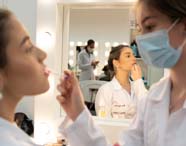 Em um laboratório de estética e cosmética, estudantes com jalecos brancos realizam atividade prática de maquiagem. Uma aluna aplica produto no rosto de outra sentada diante de espelho iluminado por lâmpadas circulares. Ao fundo, outros alunos também praticam, refletidos nos espelhos do ambiente profissional.