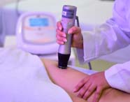 Close de uma mão segurando aparelho de radiofrequência sobre a pele da perna de uma pessoa coberta por toalha branca. O procedimento é realizado em laboratório de habilidades terapêuticas do curso de Estética e Cosmética, com equipamentos modernos ao fundo sob luz violeta.