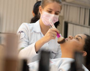 Aluna do curso de Estética e Cosmética, usando jaleco branco com símbolo do curso na manga, aplica maquiagem com pincel rosa no rosto de uma colega deitada. Frascos de produtos aparecem desfocados em primeiro plano, sugerindo ambiente de prática em sala de aula.