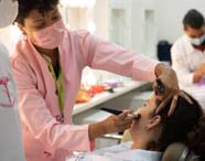 Ambiente de laboratório de estética com várias estudantes em jalecos brancos. Uma instrutora de jaleco rosa e crachá verde aplica maquiagem em uma aluna sentada, enquanto outras duplas treinam técnicas semelhantes ao fundo, cercadas por espelhos iluminados e bancadas com materiais de beleza