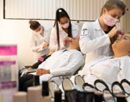 Sala de aula prática do curso de Estética e Cosmética com várias alunas em jalecos brancos aplicando maquiagem umas nas outras. Elas estão sentadas em cadeiras diante de espelhos iluminados, com potes de pó compacto e outros itens de maquiagem organizados sobre a bancada.
