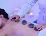 Estudante de Estética e Cosmética realiza massagem com pedras quentes, posicionando cuidadosamente pedras lisas e escuras ao longo das costas de uma pessoa deitada sobre maca, em ambiente de iluminação suave e relaxante, com vela acesa ao fundo.