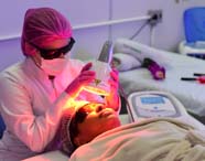Profissional ou estudante de Estética e Cosmética, com touca rosa e jaleco branco, segura aparelho de fototerapia emitindo luz vermelha sobre o rosto de paciente protegido por óculos escuros, em maca coberta por manta bege, dentro de laboratório equipado com aparelhos modernos.