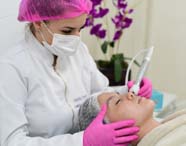 Aluna do curso de Estética e Cosmética, usando jaleco branco e touca rosa, realiza procedimento facial com aparelho moderno em paciente deitada, que usa touca descartável. Ao fundo, flores roxas decoram o ambiente, indicando cuidado e atenção ao bem-estar.