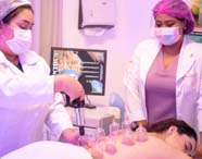 Duas mulheres em jalecos brancos, uma com touca, aplicam ventosas transparentes nas costas de paciente deitada em maca. A cena ocorre em laboratório de Estética e Cosmética, com monitor ligado ao fundo exibindo informações sobre procedimentos terapêuticos.