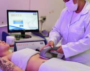 Estudante de Estética e Cosmética realiza procedimento corporal utilizando aparelho grande sobre abdômen de paciente tatuada, deitada em maca coberta por toalha branca. Monitor de computador exibe tela do equipamento Hecus, ambiente limpo e bem iluminado.