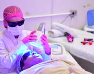 Estudante de Estética e Cosmética realiza procedimento corporal utilizando aparelho grande sobre abdômen de paciente tatuada, deitada em maca coberta por toalha branca. Monitor de computador exibe tela do equipamento Hecus, ambiente limpo e bem iluminado.