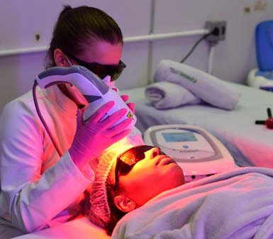 Profissional de Estética e Cosmética realiza procedimento facial com aparelho de luz pulsada em paciente deitada, que usa touca descartável e óculos de proteção. O ambiente é clínico, com lençóis brancos, toalhas enroladas e equipamentos modernos, típico de laboratório universitário da área.