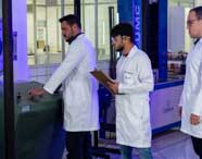 Estudantes de Engenharia Civil em laboratório, vestindo jalecos brancos, operam máquina de ensaio mecânico. Um aluno anota dados em prancheta, enquanto outro manuseia o equipamento. Ao fundo, há janelas e máquinas industriais, ambiente típico de laboratório universitário.