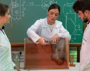 Professora de Engenharia Civil demonstra experimento sobre solos utilizando caixa transparente com terra, enquanto dois alunos assistem atentos. No quadro verde ao fundo, diagramas técnicos ilustram conceitos de mecânica dos solos, caracterizando aula prática universitária.