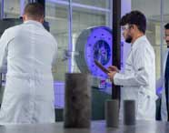 Estudantes de Engenharia Civil, de costas, analisam equipamento de teste de resistência de materiais. Usam jalecos brancos e seguram pranchetas para anotações. O laboratório possui máquinas robustas, instrumentos de medição e iluminação azulada, típico de ambientes acadêmicos.