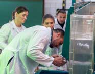 Grupo de estudantes e professor de Engenharia Civil, todos de jaleco branco, observam atentamente experimento em tanque transparente com fluxo de água. O professor se inclina para ajustar o equipamento, enquanto alunos acompanham a atividade prática no laboratório.