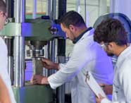 Professor de Engenharia Civil orienta alunos durante teste de compressão em prensa hidráulica. Todos usam jaleco branco; um aluno segura prancheta para registrar dados. O ambiente é repleto de equipamentos industriais, evidenciando prática laboratorial universitária.