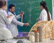 Três estudantes de Engenharia Civil, dois homens e uma mulher, observam atentamente experimento em máquina de testes. O grupo usa jalecos brancos e está posicionado próximo ao equipamento, acompanhando instruções técnicas do professor em ambiente de laboratório.