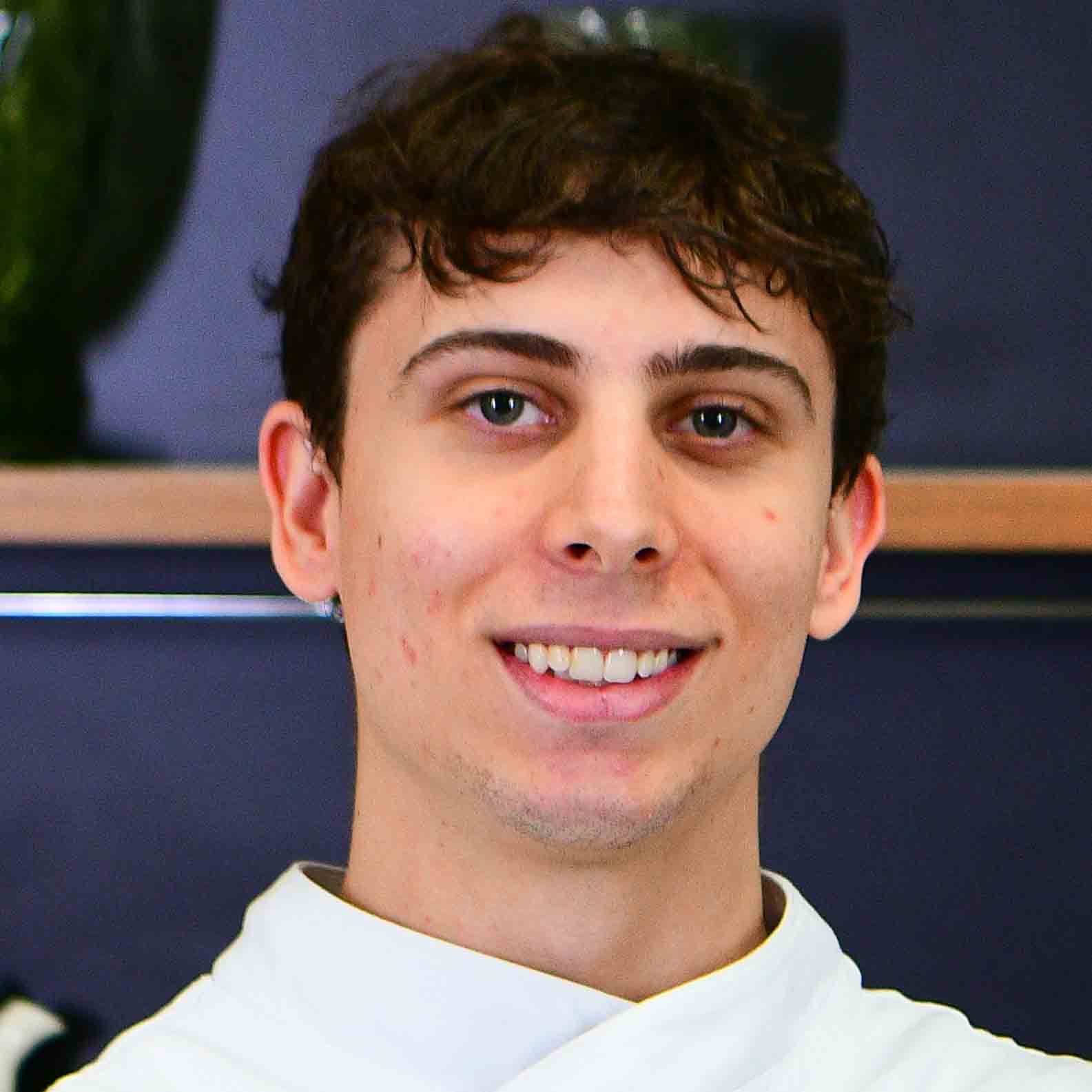Allan, aluno do curso de gastronomia, trajando um uniforme branco de chef. Ele está em um ambiente interno com fundo escuro decorado por prateleiras e objetos, sugerindo uma cozinha ou espaço gastronômico profissional. A iluminação clara destaca o contraste entre o uniforme e o fundo, reforçando o contexto culinário da imagem.