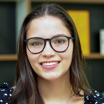 Maria, aluna do curso de gastronomia, trajando uma blusa preta com detalhes brancos. Ela está em um ambiente interno com fundo composto por estantes e livros, sugerindo um espaço de estudo ou biblioteca. A iluminação suave valoriza os cabelos castanhos e o óculos, criando um contraste harmonioso com o cenário e reforçando o ambiente acadêmico.