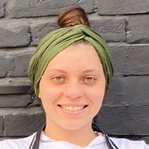 Maria Júlia, egressa do curso de gastronomia, trajando um avental cinza sobre um uniforme branco de chef e usando um lenço verde na cabeça. Ela está em um ambiente externo com fundo de parede cinza texturizada, sugerindo um espaço urbano ou restaurante ao ar livre. A iluminação natural destaca os elementos do uniforme e transmite um ar descontraído e profissional.