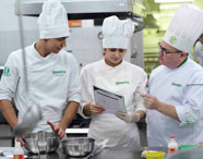 Três pessoas com domas brancas e toques de chef, todos com o logo da Unoeste, estão em uma cozinha profissional. Uma delas segura um menu ou ficha técnica enquanto outra aponta para o papel, sugerindo orientação prática entre professor e estudantes do curso de Gastronomia da Unoeste. Na bancada, há tigelas metálicas e utensílios culinários.