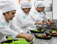 Três estudantes do curso de Gastronomia da Unoeste, com domas e toques brancos, preparam saladas em recipientes pretos sobre bancada de granito. Cada um monta porções individuais com folhas verdes e ingredientes coloridos, demonstrando trabalho em equipe na cozinha didática.