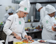 Estudantes de Gastronomia da Unoeste, vestindo domas e toques brancos, trabalham em uma cozinha profissional. Em primeiro plano, uma aluna com tatuagem no antebraço corta alimentos sobre tábua, enquanto outros colegas cozinham e organizam ingredientes ao fundo, ilustrando atividade prática do curso.