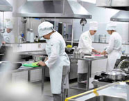 Ambiente amplo de cozinha industrial com vários estudantes de Gastronomia da Unoeste, todos uniformizados, realizando diferentes tarefas culinárias. Uma aluna corta vegetais em tábua verde, enquanto outros manuseiam ingredientes e utensílios nas bancadas de inox, evidenciando dinâmica de aula prática.