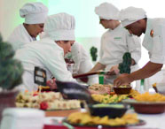 Grupo de estudantes do curso de Gastronomia da Unoeste, uniformizados com doma e toque de chef, monta uma mesa repleta de pratos coloridos e petiscos variados. Os alunos organizam e finalizam os preparos, demonstrando integração e colaboração em evento gastronômico institucional.