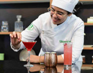 Estudante de Gastronomia da Unoeste prepara drinks em bancada. À frente, três bebidas: um coquetel vermelho em taça martini, um drink claro em caneca de cobre e outro rosado em copo alto com cereja. O cenário inclui prateleiras com potes e utensílios, sugerindo aula de mixologia.