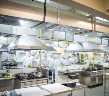 Cozinha didática moderna e espaçosa, equipada com fogões industriais, coifas de aço inox, bancadas amplas e utensílios culinários. Estudantes do curso de Gastronomia, todos com uniformes brancos e toques, reúnem-se em grupo para atividade prática na instituição de ensino.