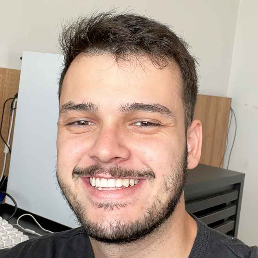 Leonardo, egresso de Marketing, posando em um ambiente interno com uma mesa e um computador ao fundo. Ele tem barba e um sorriso largo.