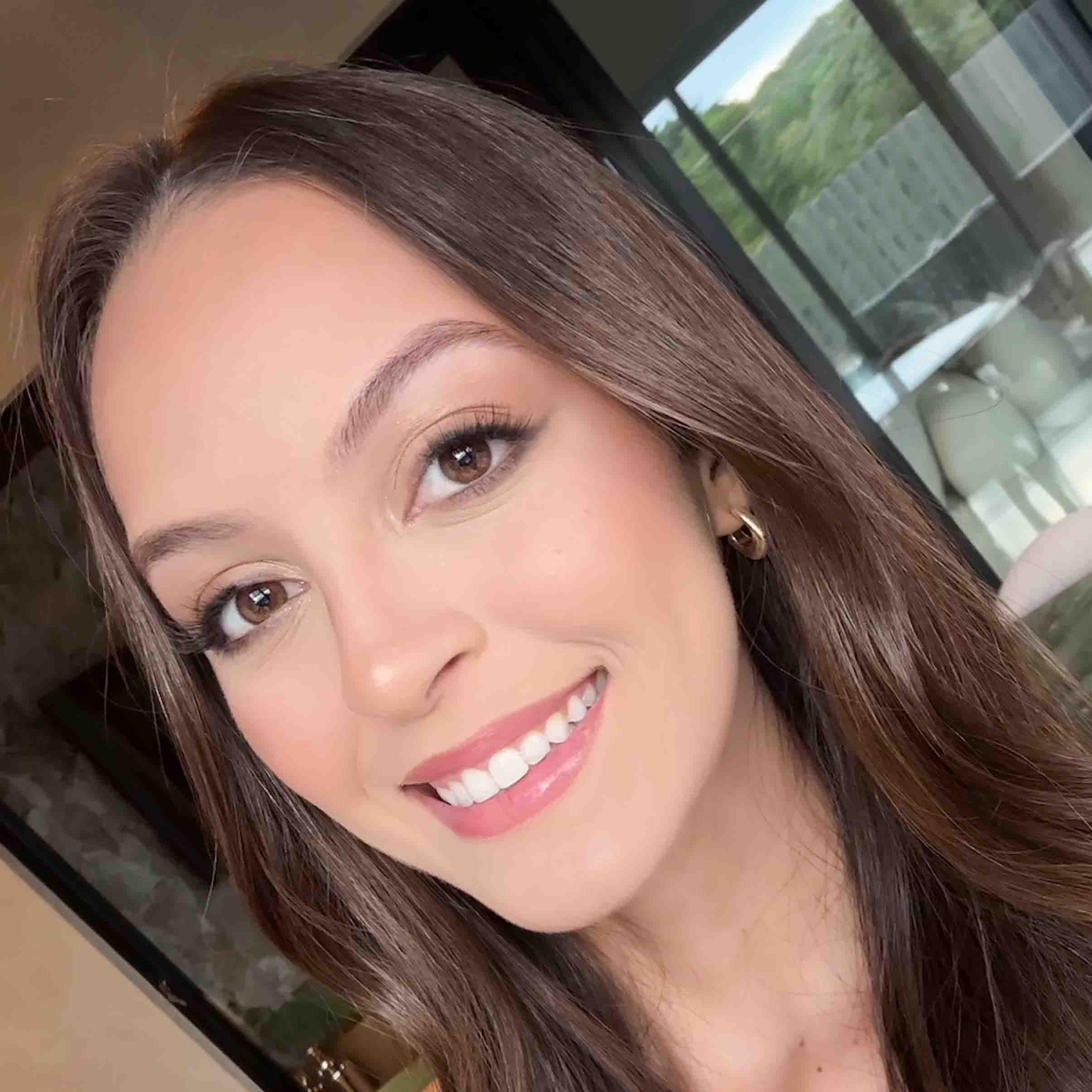 Raiane, aluna de Marketing, posando em um ambiente interno com uma janela ao fundo. Ela usa brincos de argola dourados e tem cabelos longos e lisos, com um sorriso largo.
