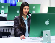 Estudante do curso de Marketing sentada em laboratório de informática, utilizando computador iMac verde. Ela veste camisa azul e jaqueta escura, com cabelos longos e lisos. Ao fundo, outras estações de trabalho organizadas em fileiras, ambiente claro e moderno da universidade.