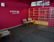 Espaço interno da Business School com parede vermelha, logo institucional e prateleiras de madeira. Duas poltronas cinza-claro estão dispostas no canto, ao lado de uma divisória metálica. Ambiente acolhedor, projetado para reuniões, estudos ou espera dos estudantes de Marketing.