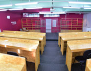 Auditório da Business School com carteiras de madeira clara organizadas em fileiras, piso escuro e paredes vermelhas. Ao fundo, estantes vazias, cadeiras de espera e divisória metálica. Espaço amplo, iluminado, destinado a aulas, palestras e eventos acadêmicos do curso de Marketing.