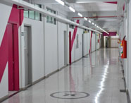 Corredor amplo e bem iluminado, com paredes brancas e detalhes em vermelho, portas de salas de aula alinhadas à esquerda. Piso claro e brilhante, extintores de incêndio fixados nas paredes, ambiente organizado e seguro, típico das instalações da faculdade de Marketing.