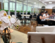 Sala ampla com paredes revestidas de madeira, mesas redondas e cadeiras pretas ocupadas por estudantes em atividades de grupo. Vaso de orquídea branca em primeiro plano, iluminação suave e ambiente acolhedor, proporcionando espaço de convivência e estudo para alunos de Marketing.