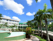 Vista externa do bloco B3 da Unoeste, com edifícios de vários andares, palmeiras altas e jardins bem cuidados. Céu azul com nuvens brancas, fonte de água e passarelas verdes compõem o cenário, destacando a infraestrutura moderna e arborizada da instituição.
