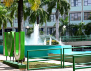 Fonte central no campus da Unoeste, rodeada por palmeiras e gramado. Em destaque, escultura verde “I love U” próxima à fonte com jatos d’água. Ao fundo, prédio da universidade com várias janelas, compondo um ambiente vibrante e acolhedor para os estudantes de Marketing.