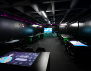 Sala de aula escura e moderna, equipada com mesas digitais interativas, cadeiras giratórias e iluminação indireta roxa. Ao fundo, grande tela digital para apresentações. Espaço inovador voltado para dinâmicas, simulações e metodologias ativas no ensino de Marketing.