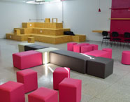 Sala multifuncional com arquibancada de madeira clara, pufes coloridos em tons de rosa, cinza e branco espalhados pelo chão. À direita, bancada com cadeiras e luminárias vermelhas. Ambiente versátil, ideal para aulas criativas, workshops e atividades colaborativas do curso.