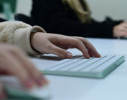 Close nas mãos de estudantes digitando em teclados de iMac verde sobre mesa branca. As mangas de roupas claras e escuras aparecem parcialmente, evidenciando concentração e prática em laboratório de informática, fundamental para as atividades do curso de Marketing.