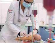 Estudante do curso de Enfermagem, com jaleco branco e máscara, realiza administração de medicamento intravenoso em braço de manequim. Na bancada há bolsa de soro vermelho, materiais médicos e estetoscópio. O ambiente é um laboratório prático da faculdade.